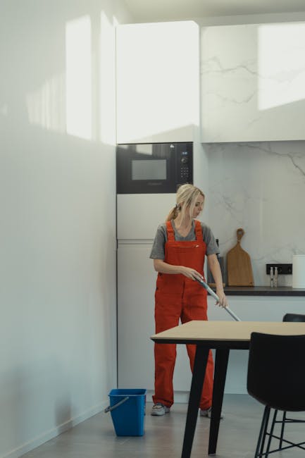 A light blue plastic dustpan and a wooden-handled scrubbing brush with natural bristles are hanging on a plain, light grey wall. The dustpan is positioned at an angle with the brush resting inside it,